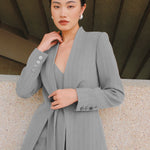 A beautiful professional model wearing an elegant grey pinstripe collarless blazer featuring a distinctive tie-front knot at the waist, styled with a crisp white collared shirt underneath for a modern corporate chic look.