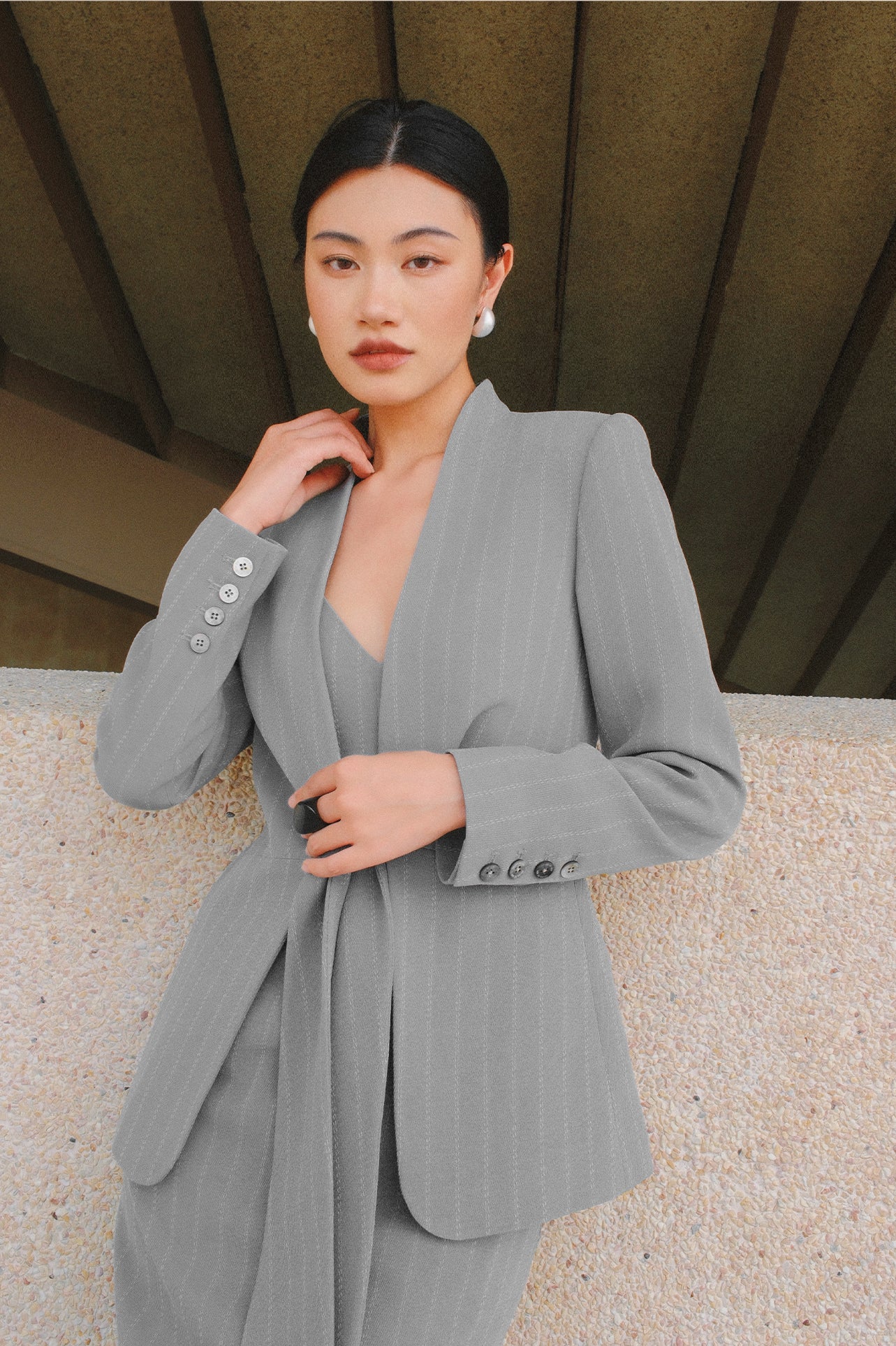A beautiful professional model wearing an elegant grey pinstripe collarless blazer featuring a distinctive tie-front knot at the waist, styled with a crisp white collared shirt underneath for a modern corporate chic look.