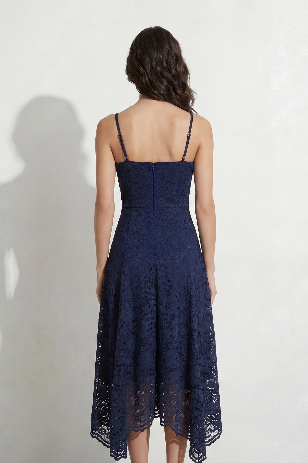 A professional woman modeling an elegant navy blue lace midi dress, featuring delicate V-neck spaghetti straps, a tailored fit-and-flare silhouette, and a beautiful flowing asymmetric handkerchief lace trim hem.
