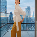 A stylish woman posing against a city skyline wearing a navy blue wrap blouse with golden ginkgo leaf print tied at the waist, paired with tailored mustard yellow wide-leg trousers.