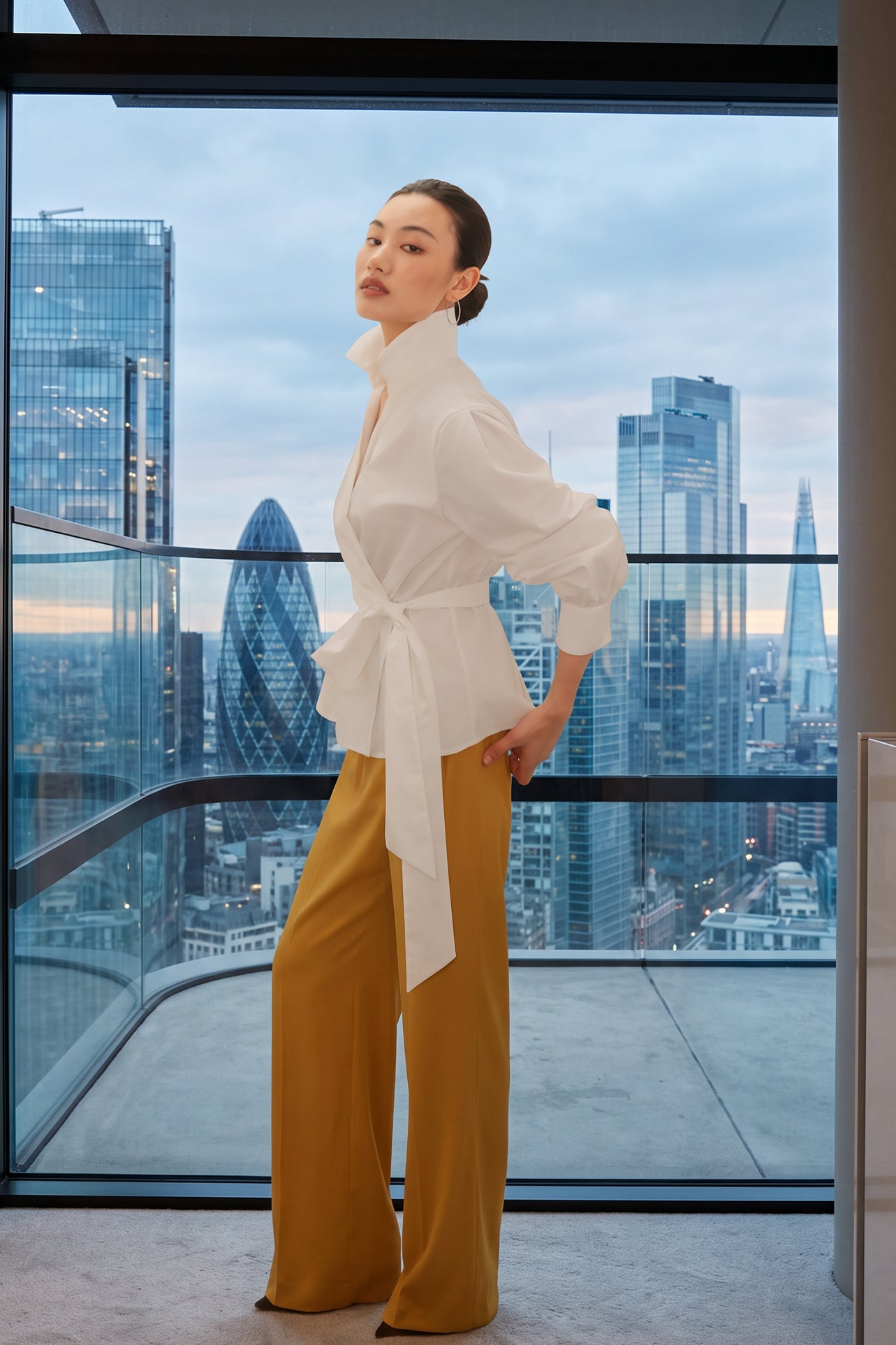 A stylish woman posing against a city skyline wearing a navy blue wrap blouse with golden ginkgo leaf print tied at the waist, paired with tailored mustard yellow wide-leg trousers.