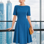 A beautiful model wearing a sophisticated medium blue A-line midi dress featuring an elegant boat neckline with refined asymmetric shoulder pleating, short sleeves, and a flowing flared swing skirt.