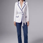 An elegant professional woman wearing a tailored white blazer with navy blue contrast piping, featuring a self-tie belt to define the waist for a smart-casual office look