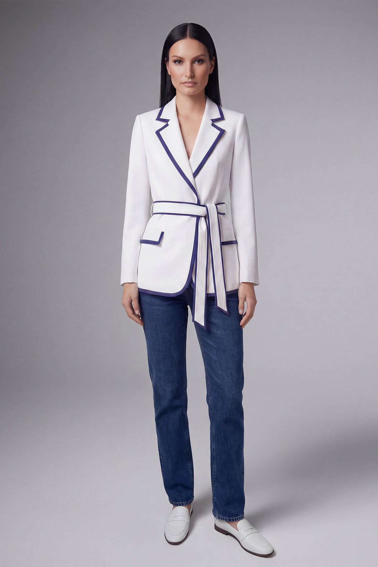 An elegant professional woman wearing a tailored white blazer with navy blue contrast piping, featuring a self-tie belt to define the waist for a smart-casual office look